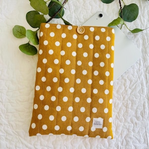 May include: A mustard yellow quilted tablet sleeve with white polka dots. The sleeve has a button closure and a small white label with the text "Lola & Grace" on it.