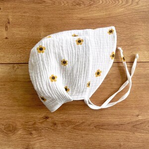 May include: White baby bonnet with a repeating pattern of yellow and brown sunflowers. The bonnet is made of a textured fabric and has white ties. The bonnet is laying on a wooden surface.