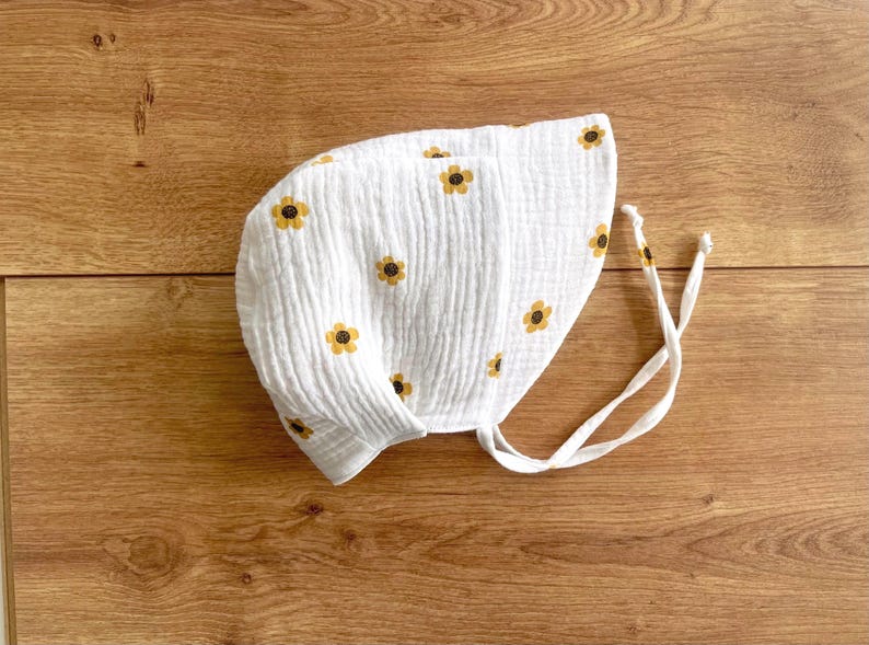 May include: White baby bonnet with a repeating pattern of yellow and brown sunflowers. The bonnet is made of a textured fabric and has white ties. The bonnet is laying on a wooden surface.
