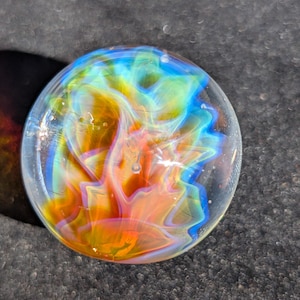 May include: A clear glass marble with vibrant, swirling colors of orange, yellow, blue, and green. The internal design resembles a nebula or a stylized flower, creating a unique and artistic appearance.