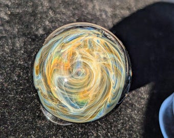 May include: A clear glass marble with an internal design of swirling gold, blue, and white colors. The marble is round and sits on a dark, textured surface. The design resembles a galaxy or nebula.
