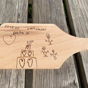 May include: A light-colored wooden cutting board with a handle. The board has a heart, a cake, and stick figures etched into the wood. The words "JOYEUX ANNIVERSAIRE PAPA" are written above the heart.