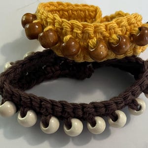 May include: Two handmade crochet bracelets. One is yellow with brown wooden beads, the other is dark brown with white beads. The bracelets are made with a thick yarn and have a textured appearance. The beads are evenly spaced around the bracelets.