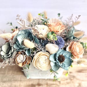 May include: A floral arrangement in a white square container. The arrangement features a variety of flowers in shades of blue, cream, and peach, with accents of dried grasses and greenery. A starfish and seashells are also included.