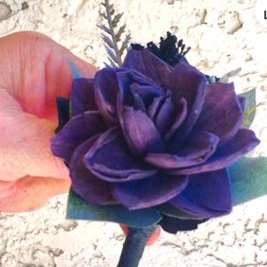 Peut inclure: Une boutonni&egrave;re florale violet fonc&eacute; avec des p&eacute;tales superpos&eacute;s, accentu&eacute;e par de petites feuilles vertes et une tige sombre. La fleur est tenue dans une main sur un fond blanc textur&eacute;. Une petite brindille de verdure ajoute des d&eacute;tails.