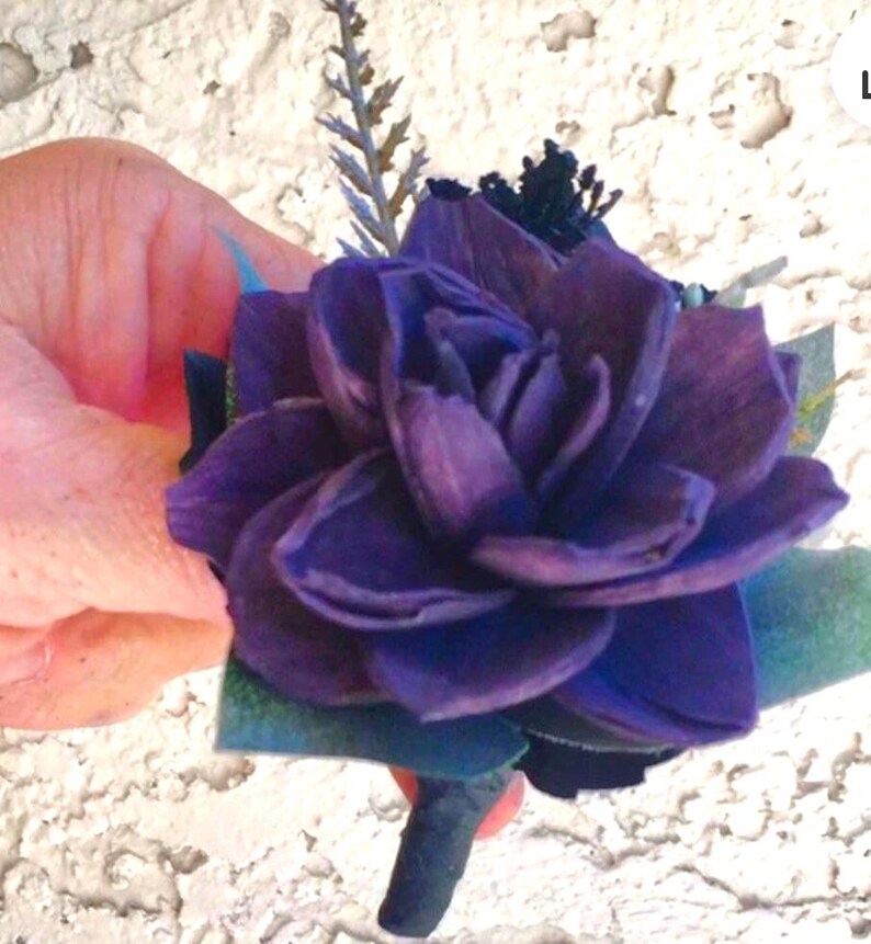 Peut inclure: Une boutonni&egrave;re florale violet fonc&eacute; avec des p&eacute;tales superpos&eacute;s, accentu&eacute;e par de petites feuilles vertes et une tige sombre. La fleur est tenue dans une main sur un fond blanc textur&eacute;. Une petite brindille de verdure ajoute des d&eacute;tails.