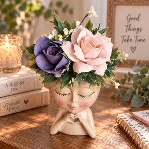 May include: A beige resin planter shaped like a human head with closed eyes and hands forming a heart shape. The planter holds a floral arrangement featuring one large purple rose, one large light pink rose, green leaves, and small white buds.