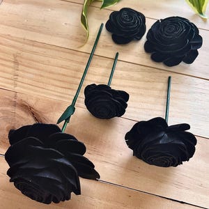 May include: An assortment of black artificial roses with green stems and leaves. The roses vary in size, with some fully bloomed and others in bud form. The flowers are arranged on a light brown wooden surface.