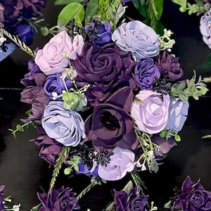 Peut inclure: Un bouquet de fleurs artificielles dans des tons de violet, de lavande et de rose clair. L'arrangement pr&eacute;sente diff&eacute;rents types de fleurs, dont des roses et de plus petites fleurs, avec des accents de feuillage vert. Le bouquet est pos&eacute; sur un fond sombre.