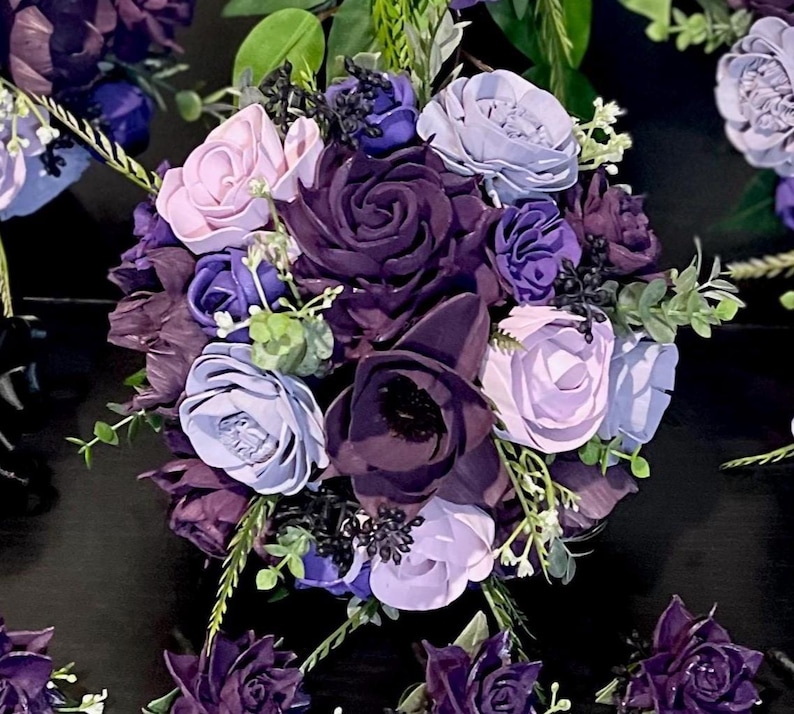 Peut inclure: Un bouquet de fleurs artificielles dans des tons de violet, de lavande et de rose clair. L'arrangement pr&eacute;sente diff&eacute;rents types de fleurs, dont des roses et de plus petites fleurs, avec des accents de feuillage vert. Le bouquet est pos&eacute; sur un fond sombre.