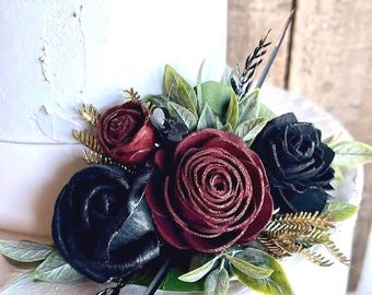 May include: A floral arrangement featuring deep burgundy and black roses, accented with green leaves and gold-toned foliage. The flowers are clustered together, displaying layered petals and a dark, rich color palette against a white surface.