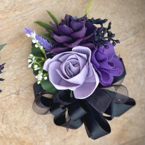 Peut inclure: Une boutonni&egrave;re florale artisanale avec des roses violettes et lavande, agr&eacute;ment&eacute;e de petites fleurs blanches, de feuilles vertes et d'un n&oelig;ud en ruban noir. L'arrangement est con&ccedil;u pour les occasions sp&eacute;ciales.