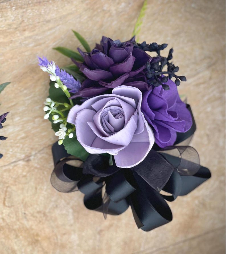 Peut inclure: Une boutonni&egrave;re florale artisanale avec des roses violettes et lavande, agr&eacute;ment&eacute;e de petites fleurs blanches, de feuilles vertes et d'un n&oelig;ud en ruban noir. L'arrangement est con&ccedil;u pour les occasions sp&eacute;ciales.