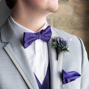 May include: A gray suit with a white dress shirt, a purple bow tie, and a matching pocket square. A purple floral boutonniere is pinned to the lapel. The suit jacket has a purple patterned vest underneath.