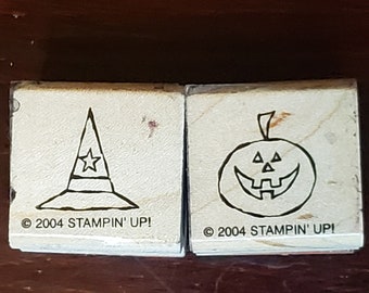 May include: Two wooden stamps with black line drawings. The left stamp shows a witch's hat with a star on top. The right stamp shows a jack-o'-lantern with a wide grin. Both stamps have the text "© 2004 STAMPIN' UP!" below the image.