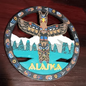Könnte beinhalten: Ein runder, schwarzer Holzschmuck mit einem bunten Bild eines indianischen Totempfols mit einem Vogel obenauf. Der Totempfahl steht vor einer schneebedeckten Bergkette mit grünen Bäumen. Die Worte "ALASKA" sind in Gelb auf dem Schmuckstück geschrieben.