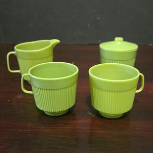 Puede incluir: Un juego de té vintage de plástico verde, que incluye dos tazas con asa, una jarra para crema y un azucarero con tapa. Las tazas y el azucarero tienen un diseño acanalado. El juego se exhibe sobre una superficie de madera oscura.
