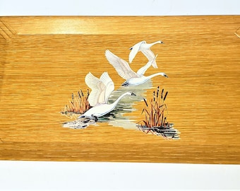 May include: A wooden serving tray featuring a painted design of three white swans in flight over a pond with reeds. The tray has a light brown wood grain and a rounded edge.