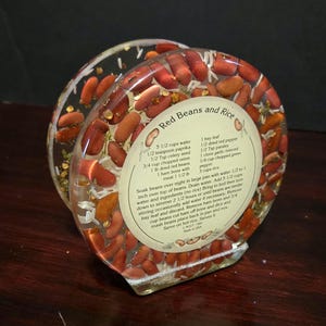 May include: A clear resin coaster with red beans and white rice embedded inside. The coaster has a recipe for "Red Beans and Rice" printed on it, including ingredients like water, paprika, celery seed, onion, dried red beans, ham bone, bay leaf, red pepper, parsley, garlic, green pepper, and rice.