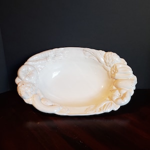 May include: White ceramic serving bowl with a raised design of vegetables around the rim. The bowl is oval shaped and has a smooth, white glaze.