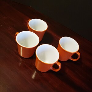 Puede incluir: Cuatro tazas de cerámica naranja con interiores blancos. Las tazas están dispuestas en forma de cuadrado sobre una superficie de madera marrón oscuro.