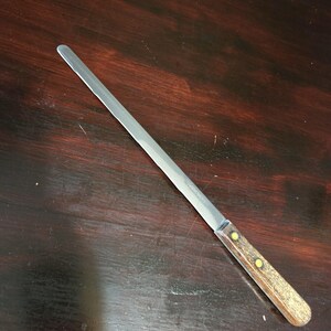 May include: A long, stainless steel bread knife with a wooden handle. The handle is light brown with two yellow rivets. The blade is thin and straight, designed for slicing bread and pastries.