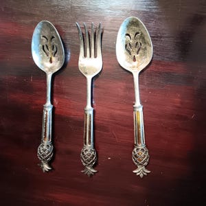 May include: Three silver utensils with pineapple-shaped handles. The utensils are a slotted spoon, a fork, and a serving spoon.