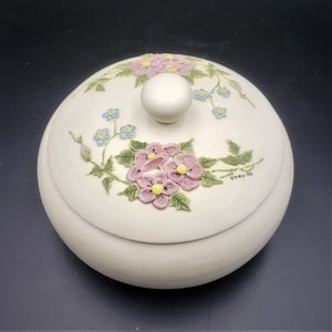 Peut inclure: Boîte à bijoux en céramique blanc cassé avec couvercle et bouton rond. Le couvercle est orné de fleurs roses et jaunes, de fleurs bleues et de feuilles vertes. La signature de l'artiste "Gwenn '92" est visible sur le couvercle.