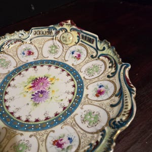 May include: An antique porcelain plate with a cream-colored background, adorned with hand-painted floral designs in shades of purple, pink, and blue. The plate features a teal blue border and gold accents, with a scalloped edge.