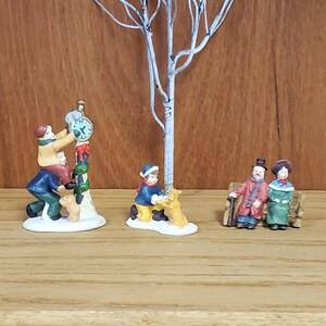 May include: A miniature Christmas village scene with three figures. One figure is a man in a blue coat and red hat, holding a small dog. Another figure is a boy in a yellow coat and blue hat, holding a dog. The third figure is a couple sitting on a bench. The scene is set in a snowy winter setting.