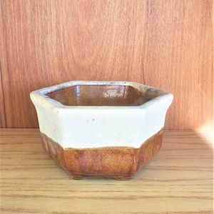 Può includere: Un piccolo vaso esagonale in ceramica con un design bicolore. La metà superiore è bianco crema, mentre la metà inferiore è marrone terroso. Il vaso ha una superficie leggermente strutturata e poggia su tre piccoli piedini.