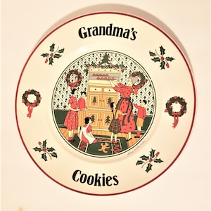Puede incluir: Un plato de cerámica blanco con un borde rojo presenta una escena navideña con una mujer horneando galletas en una cocina. El plato tiene el texto "Grandma's Cookies" y un diseño de acebo verde.