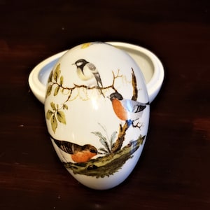 Op de afbeelding: Witte porseleinen eivormige sieradenbox met een deksel. De doos is versierd met een handgeschilderd ontwerp van drie vogels die op takken met groene bladeren zitten. De vogels zijn een zwart-witte vogel, een roodborstje en een bruin-witte vogel.