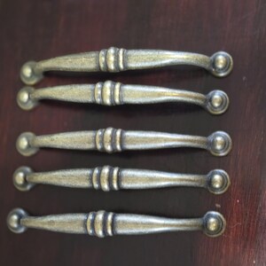 Op de afbeelding: Vijf antieke stijl kastgrepen met een geborsteld messing afwerking. Elke greep heeft aan het ene uiteinde een ronde knop en in het midden een decoratief, geribbeld ontwerp.