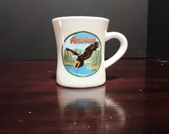 Może przedstawiać: Biały ceramiczny kubek z zakrzywionym uchwytem. Kubek posiada okrągłą grafikę z napisem "American Expedition" oraz wizerunek orła w locie nad jeziorem i lasem. Kubek ma lekko rozszerzony brzeg.