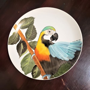 May include: A white ceramic plate featuring a hand-painted design of a parrot perched on a branch with green leaves. The parrot has yellow, orange, blue, and green plumage.