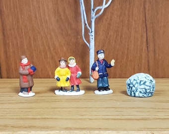 May include: A set of four miniature figurines depicting people in winter clothing. The figurines are made of painted ceramic and feature a mail carrier, two children, and a person wearing a brown coat and hat. The figurines are arranged on a wooden surface with a small, gray, textured rock in the background.