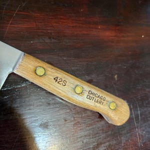 May include: Close-up of a vintage Chicago Cutlery knife. The wooden handle is light brown with three brass rivets and the text "42S" and "CHICAGO CUTLERY" etched into the wood. The blade is silver and angled.