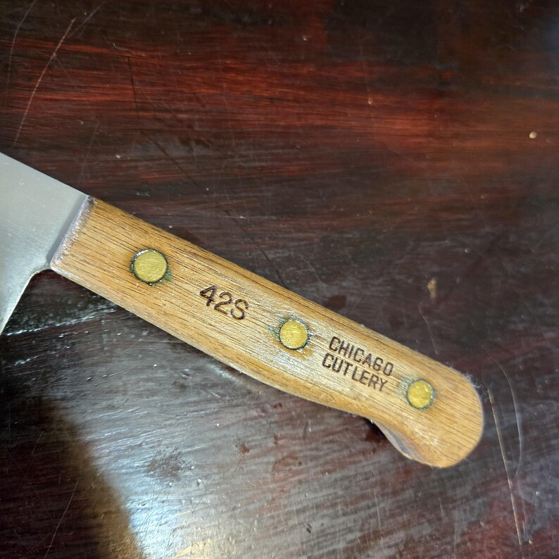 May include: Close-up of a vintage Chicago Cutlery knife. The wooden handle is light brown with three brass rivets and the text "42S" and "CHICAGO CUTLERY" etched into the wood. The blade is silver and angled.