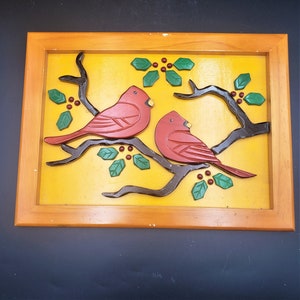 May include: A wooden framed wall art piece with a yellow background and two red cardinals perched on a branch with green leaves. The cardinals are carved and painted in a folk art style.