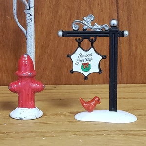 May include: A miniature Christmas sign with a black metal frame and a white star-shaped sign that reads "Season's Greetings" with a green wreath and red bow. A red cardinal bird figurine sits on a snowy base.