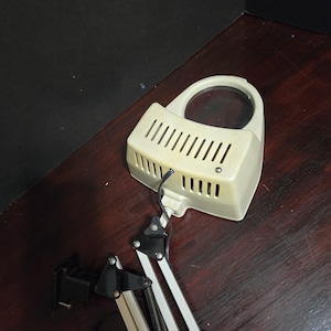 May include: A vintage off-white desk lamp with a magnifying glass and a pivoting arm. The lamp has a rectangular head with ventilation slits and a circular handle. The arm and base are black and white.