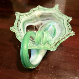 May include: A green and white glass bowl with a swirl design and a green handle. The bowl is made of art glass and has a unique, organic shape.