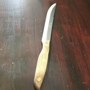 Puede incluir: Un cuchillo de cocina de acero inoxidable con mango de madera. El mango tiene una veta de madera marrón oscura y tres remaches de latón. La hoja es larga y delgada con una ligera curva.
