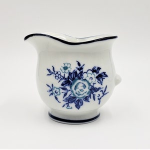 Op de afbeelding: Een witte keramische roomkan met een blauw en wit bloemenmotief. De roomkan heeft een rond lichaam en een kleine tuit.