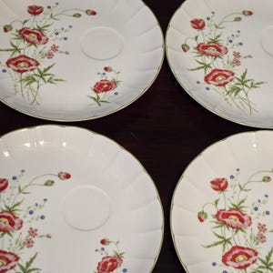 May include: Set of four white ceramic dessert plates with a floral design featuring red poppies and green leaves. The plates have a scalloped edge and a small center well.