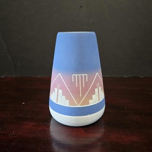 Peut inclure: Un vase en céramique bleu et blanc avec un motif géométrique. Le vase a un dessus bleu clair, une base blanche et un motif géométrique rose et blanc au milieu. Le vase est un objet décoratif.