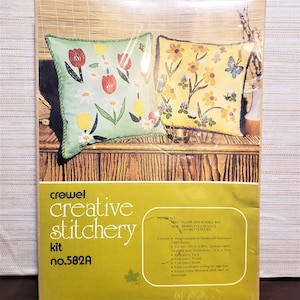 May include: A crewel embroidery kit with two pillow designs. The pillows feature floral patterns with tulips, butterflies, and bumblebees. The kit includes 14-inch by 29-inch fabric, embroidery thread, and a needle. The kit is labeled "crewel creative stitchery kit no. 582A".