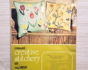 May include: A crewel embroidery kit featuring two pillow designs. The pillows depict floral patterns with tulips, butterflies, and bumblebees. The kit includes 14-inch by 29-inch fabric, embroidery thread, and a needle. The kit is labelled "crewel creative stitchery kit no. 582A".