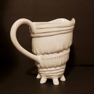 May include: White ceramic pitcher with a textured, shell-like design and a handle. The pitcher has four legs and a wide, rounded opening.